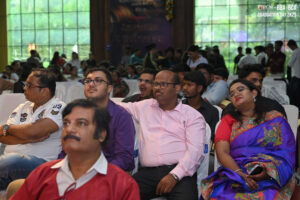 Group of formally dressed people seated at indoor graduation ceremony with large window backdrop.