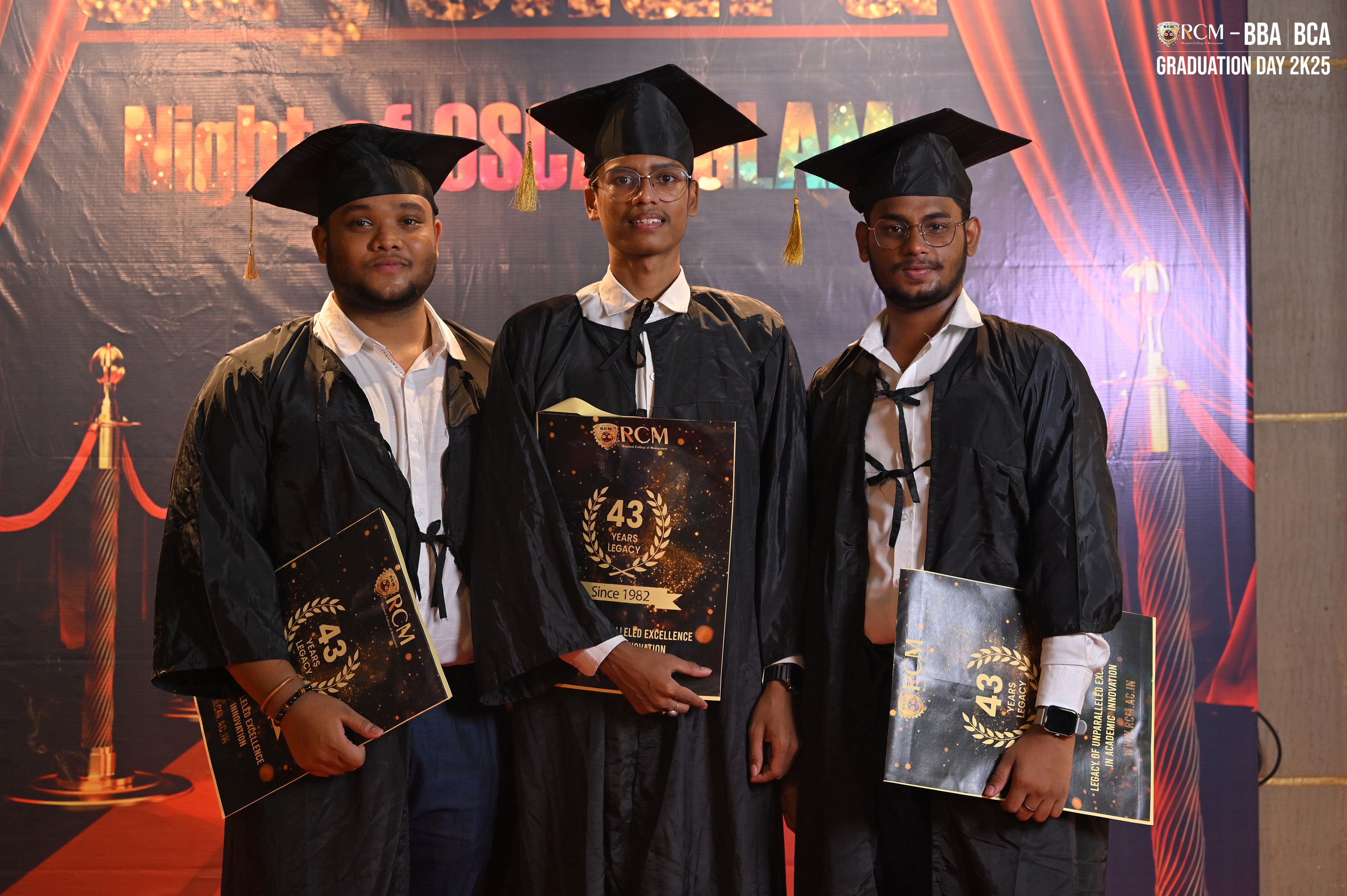 Three graduates in caps and gowns hold "RCM" folders at BBA BCA Graduation Day 2023.