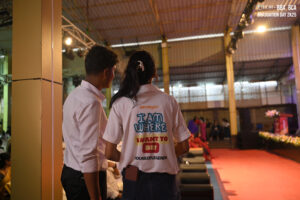 Two people stand on stage at an indoor graduation event, one wearing a shirt with motivational text.