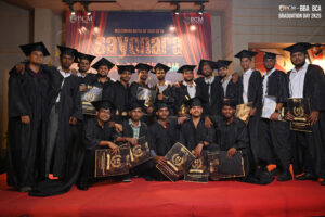 Graduates in caps and gowns holding certificates pose on red carpet at RCM BBA BCA event.