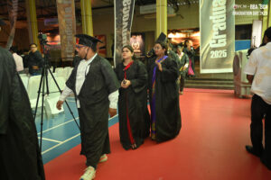 People in caps and gowns walking at a 2025 graduation ceremony with banners and red carpet.