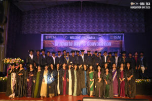 Graduates in caps and gowns on stage during RCM convocation, with banner and flowers.