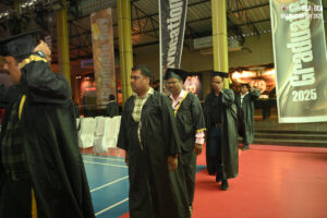 Adults in academic gowns walk in line at an indoor 2025 graduation ceremony.