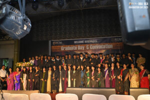 Graduates on stage at RCM Convocation, in gowns and caps, celebrating with flowers.