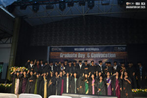 Graduates in black gowns and caps pose on stage during a "Graduate Day & Convocation" ceremony.
