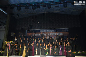 Graduates in black gowns celebrate on stage, tossing caps; RCM Global School Graduation 2025.