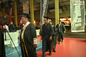 Graduates in procession indoors with banners reading "Graduation 2025" and arranged seating.