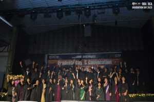 Graduates in caps and gowns celebrate on stage, tossing caps, beneath "Graduate Day & Convocation" banner.