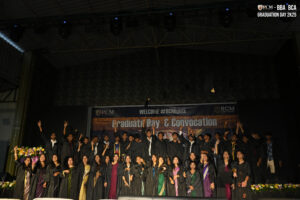 Graduates celebrate on stage in black gowns at a convocation with a "Graduate Day" banner.