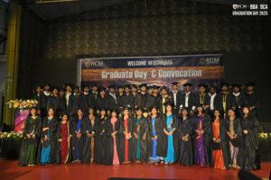 Graduates in caps and gowns on stage at "Graduate Day & Convocation" ceremony with flowers.