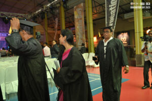 Graduates in gowns walk indoors with banners and chairs for the ceremony in the background.