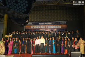 Graduates in caps and gowns at RCM Global School convocation with decorative banner background.