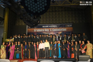 Graduates in gowns and faculty in colorful attire pose under a banner at a convocation ceremony.