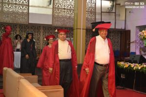 People in red academic regalia walking in indoor graduation ceremony procession with decorations.