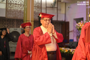 Person in red graduation gown with respectful gesture, others in similar attire, floral decor.