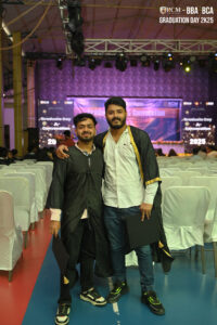 Two people in academic gowns at a 2025 graduation event in a decorated hall.