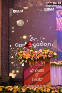 A person in red graduation attire speaks at a podium with flowers and a "42 Years of Legacy" banner.