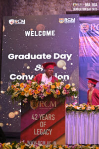 A graduate in red gown speaks at a podium with flowers, "42 Years of Legacy" backdrop.
