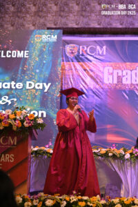 Person in red graduation gown speaking at podium during RCM Global School event.