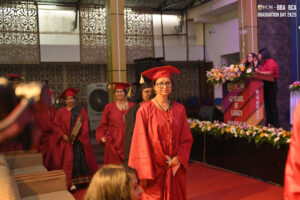 Graduates in red caps and gowns walk at a ceremony with flowers and a podium in the background.