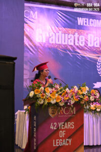 Individual in red cap and gown at RCM Graduation Day 2025 podium with floral decoration.