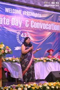 Woman in sari speaking on stage at Graduate Day & Convocation event with banner and flowers.