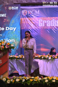 Woman speaking at RCM graduation event with flowers and banners in the background.