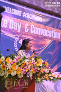Person speaks at RCM podium with flowers during 2025 BBA/BCA graduation event.