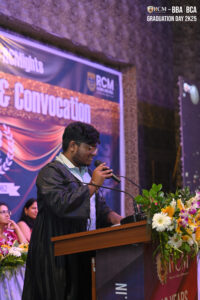 Person in graduation attire speaks at lectern during RCM Graduation Day 2K25 ceremony.
