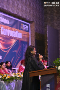 Woman in black gown speaks at podium during 2K25 convocation ceremony with banner in background.