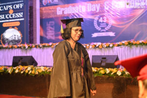 Individual in cap and gown at graduation ceremony with event banners and floral decorations.