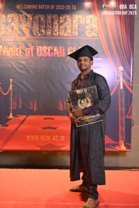 Graduate in cap and gown on red carpet, holding sign for 43rd anniversary BBA/BCA event at RCM.