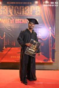 Person in graduation attire holding sign, red carpet backdrop with graduation banner text.