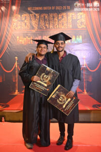 Two graduates in caps and gowns holding certificates at an RCM 2025 event backdrop.