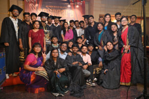 Group of graduates in caps and gowns at an indoor ceremony with welcoming backdrop.