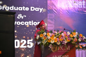 Person in red graduation gown speaks at podium with flowers, backdrop shows "Graduate Day 2025".