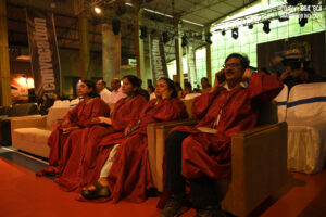 Individuals in red graduation gowns seated at an indoor convocation ceremony with banners.