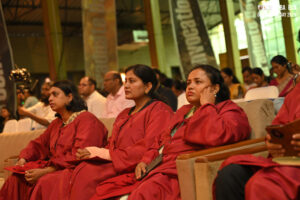Group in academic robes seated at indoor event, attentively engaged, festive banners visible.