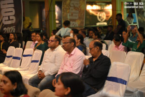 Audience seated on white chairs with blue sashes at indoor 2025 graduation event.