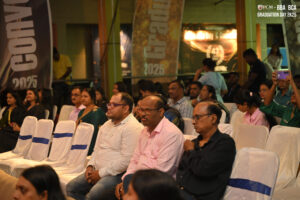 People seated at a 2025 graduation event, some taking photos, chairs with white and blue covers.