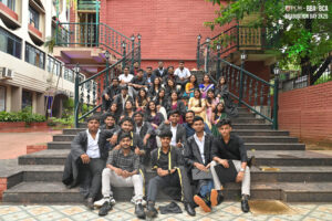 Group on stairs in formal attire for RCM BBA-BCA Graduation Day 2k23, with brick building.