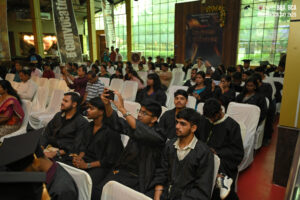 Graduation ceremony indoors, students in black gowns seated, banners "Convocation 2025".