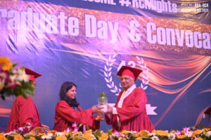 Graduates on stage, one giving a potted plant to the other, with a "Graduate Day" banner behind.