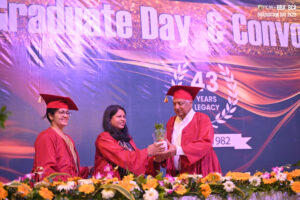 Graduation ceremony with three in academic gowns, flowers, and a banner marking 43-year legacy.