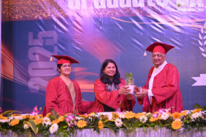 Graduates in red gowns stand with a potted plant, backdrop reads "Graduation Day 2K25".