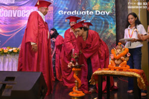 Individuals in red graduation robes light a ceremonial lamp by a Ganesha statue on Graduate Day.
