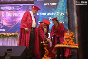 Three individuals in red robes light a ceremonial lamp on stage for Graduate Day & Convocation.