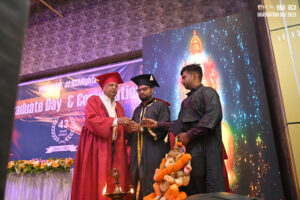 Graduation ceremony indoors, red and black gowns, colorful backdrop, decorative table.