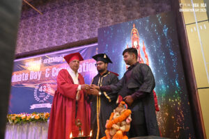 Graduation ceremony with people in academic gowns, decorative backdrop, and floral arrangement.