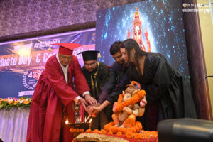 Individuals in academic robes light a ceremonial lamp near a decorated Ganesha idol.